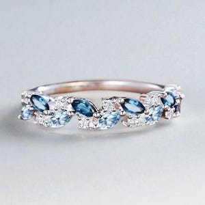 Op de afbeelding: Een zilveren ring met een herhalend patroon van marquise-geslepen blauwe edelstenen en kleine witte stenen. Het ontwerp van de ring is geplaatst tegen een zachte, neutrale achtergrond, die de glans en details van de sieraden benadrukt.