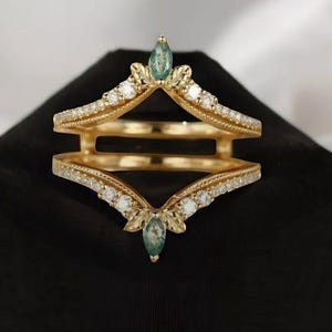 May include: A gold-colored ring guard with a double-band design. The ring features small white gemstones and two teardrop-shaped green gemstones. The ring is displayed on a black velvet surface.