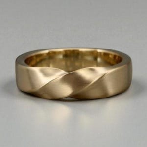 Peut inclure: Une bague dorée avec un motif torsadé. La bague a une finition mate et une bande épaisse. Le design torsadé crée une apparence subtile et texturée. La bague est probablement en or ou en métal doré.