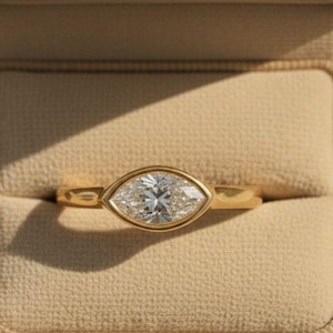 Può includere: Un anello d'oro con un diamante taglio marquise incastonato in una lunetta. L'anello è presentato all'interno di un portagioie in velluto beige. Il diamante brilla, riflettendo la luce. La fascia dell'anello è in oro lucido.