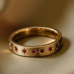 May include: A gold ring featuring a series of embedded red star-shaped gemstones. The ring is set against a soft, neutral background, highlighting the jewelry's design and colour.