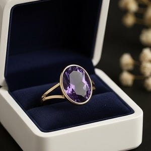 May include: A gold ring with a large oval purple gemstone, set in a white jewelry box with a navy blue interior. The ring is the focal point, showcasing the gemstone's color and shape. The box is open, displaying the ring prominently.