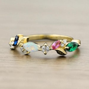 May include: A gold ring featuring a series of colorful gemstones. The ring has a central diamond-like stone, flanked by an opal, a pink stone, a green stone, and a blue stone. The gemstones are set in a leaf-like design.