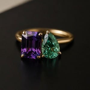 May include: A gold ring featuring two gemstones: a rectangular purple amethyst and a pear-shaped green emerald. The ring's band is a simple, polished gold, and the gemstones are set side-by-side.