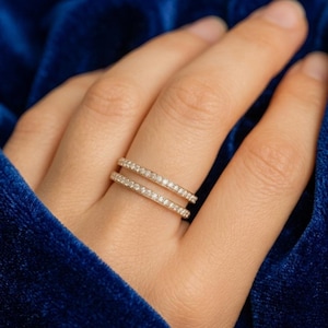 May include: A set of two thin gold bands featuring a continuous row of small, clear, sparkling stones set along the outer surface. The rings are worn stacked together on a finger, showcasing a delicate and uniform design with a polished finish.