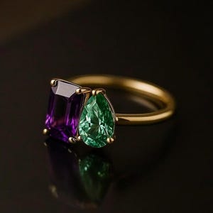 May include: A gold ring featuring a rectangular purple gemstone and a pear-shaped green gemstone. The gemstones are set side-by-side on a thin gold band. The ring is displayed against a dark background, highlighting the vibrant colors of the stones.