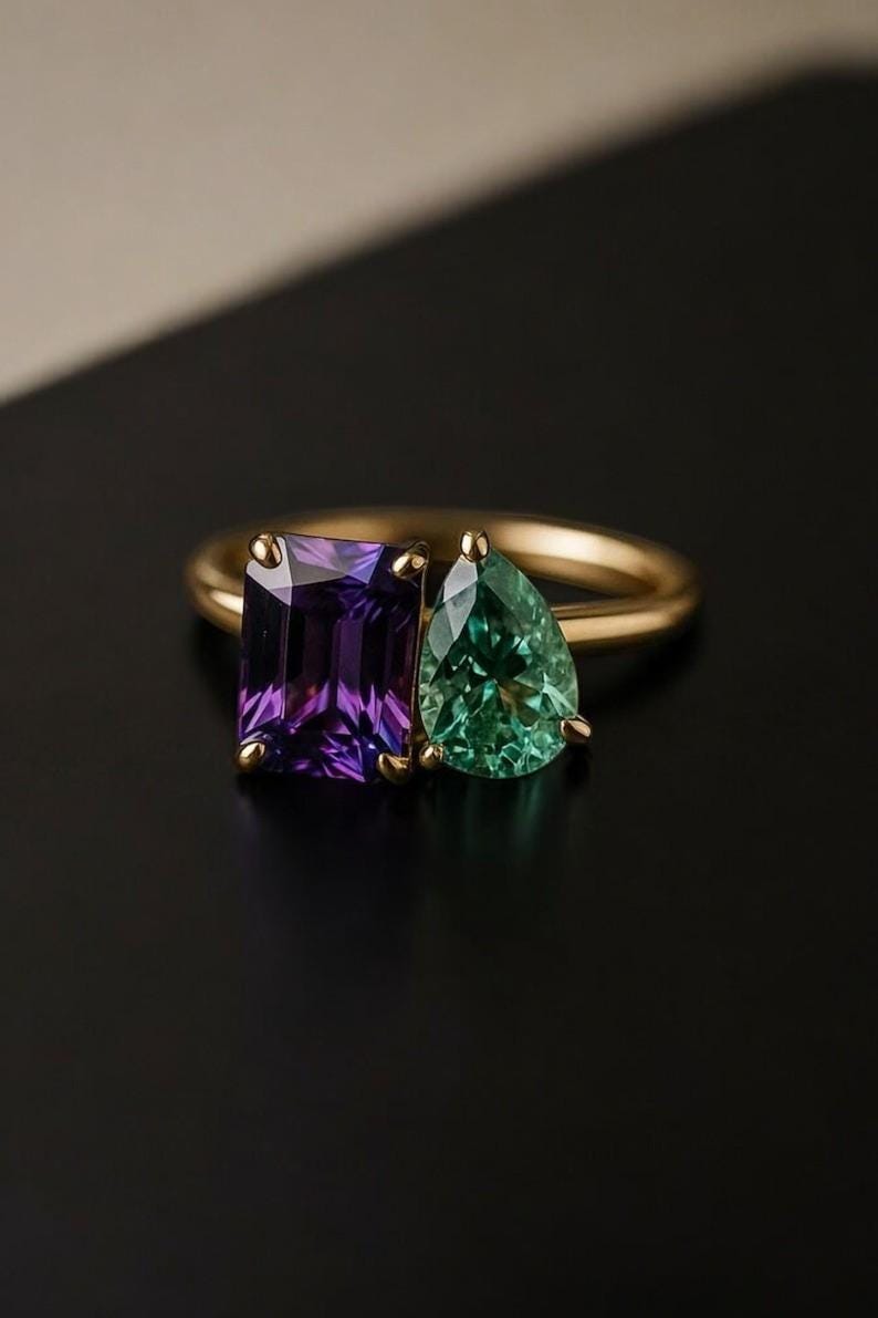 May include: A gold ring featuring two gemstones: a rectangular purple amethyst and a pear-shaped green emerald. The ring's band is a simple, polished gold, and the gemstones are set side-by-side.