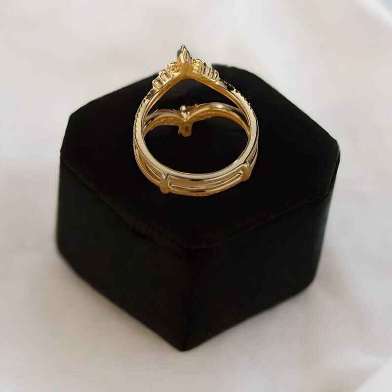 May include: A gold-colored ring with a crown design, set on a black velvet box. The ring features a double band and a pointed top with small embellishments. The ring is displayed against a white background.
