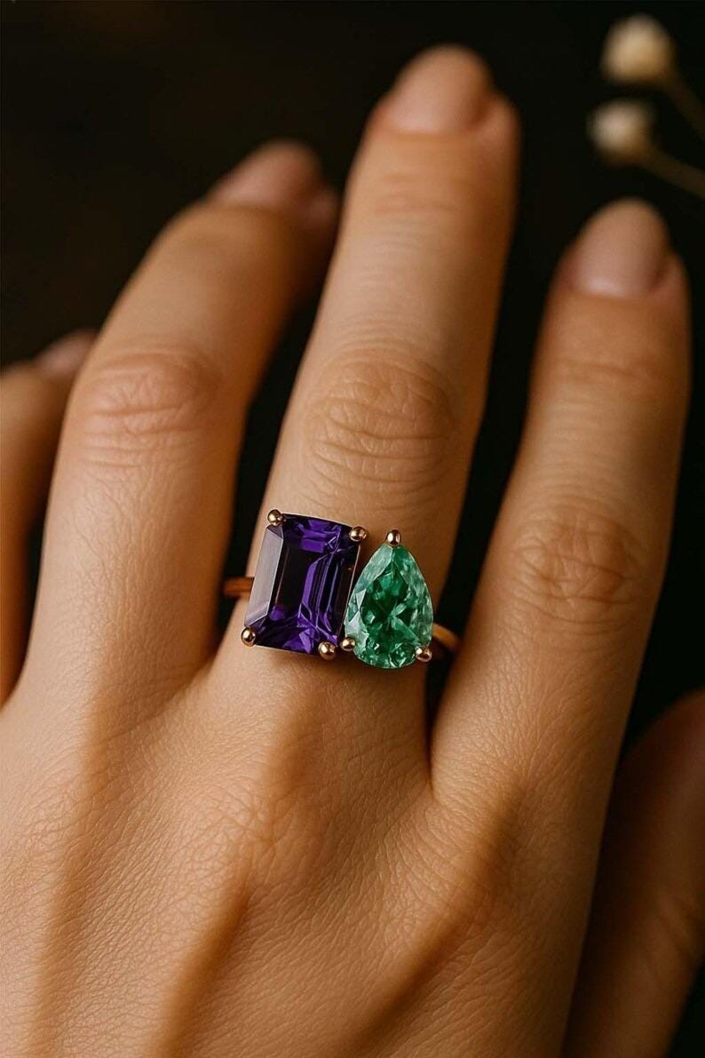 May include: A gold ring featuring two gemstones: a rectangular, deep purple amethyst and a pear-shaped, vibrant green emerald. The ring is set on a finger, showcasing the contrasting colors and shapes of the stones.