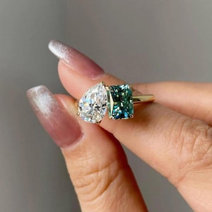 Peut inclure: Une bague en or ornée de deux pierres précieuses : une pierre claire en forme de poire et une pierre de couleur teal de taille carrée. La bague est présentée sur un fond neutre, mettant en valeur l'éclat des gemmes.