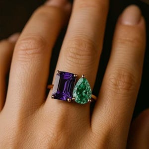 May include: A gold ring featuring two gemstones: a rectangular, deep purple amethyst and a pear-shaped, vibrant green emerald. The ring is set on a finger, showcasing the contrasting colors and shapes of the stones.