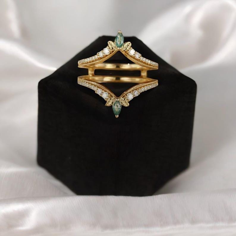 May include: A gold-colored ring guard with a double-band design. The ring features small white gemstones and two teardrop-shaped green gemstones. The ring is displayed on a black velvet surface.