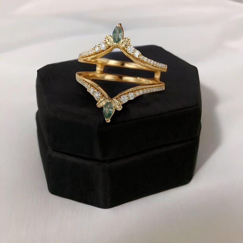May include: A gold-colored ring guard with a double band design. The ring features small, clear stones and two teardrop-shaped, green gemstones. The ring is displayed on a black, velvet-covered box against a white background.