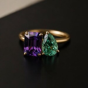 May include: A gold ring featuring two gemstones: a rectangular purple amethyst and a pear-shaped green emerald. The ring's band is a simple, polished gold, and the gemstones are set side-by-side.
