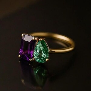 May include: A gold ring featuring a rectangular purple gemstone and a pear-shaped green gemstone. The gemstones are set side-by-side on a thin gold band. The ring is displayed against a dark background, highlighting the colors and sparkle of the gemstones.