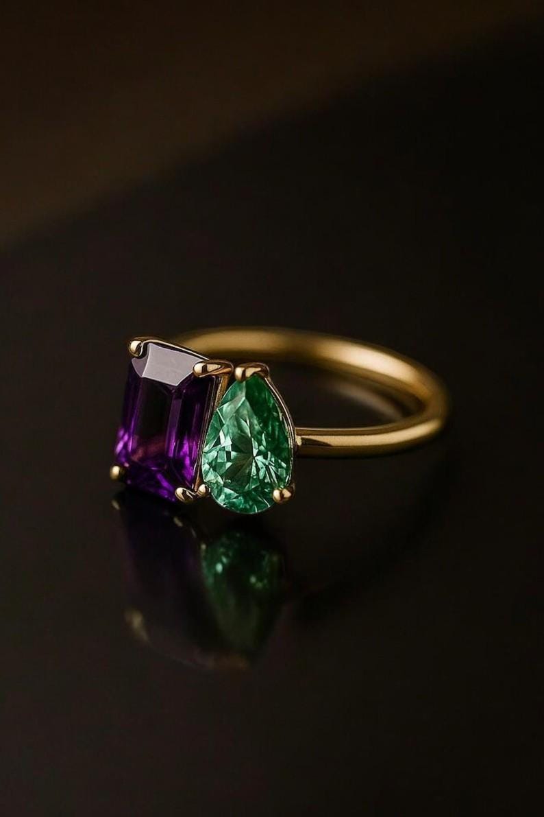 May include: A gold ring featuring a rectangular purple gemstone and a pear-shaped green gemstone. The gemstones are set side-by-side on a thin gold band. The ring is displayed against a dark background, highlighting the colors and sparkle of the gemstones.