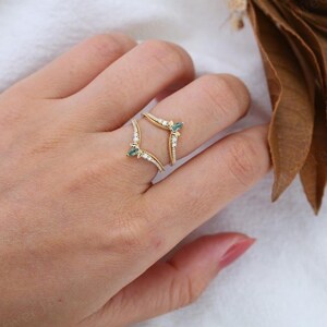 May include: A gold-colored ring with a unique design, featuring small white and teal gemstones. The ring has a double-banded structure, with the bands curving around the finger. The ring is adorned with small, sparkling white stones.