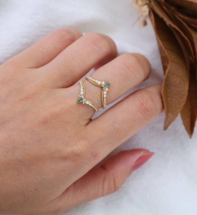 May include: A gold-colored ring with a unique design, featuring small white and teal gemstones. The ring has a double-banded structure, with the bands curving around the finger. The ring is adorned with small, sparkling white stones.