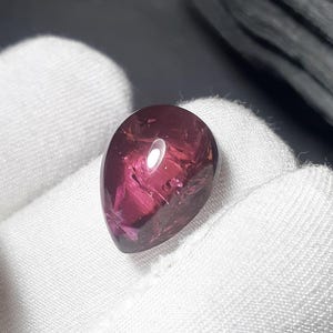 May include: A teardrop-shaped, polished gemstone with a deep magenta hue. The gem has internal inclusions and a reflective surface, held against a white fabric background. The stone's shape is smooth and rounded, with a slight point at the bottom.