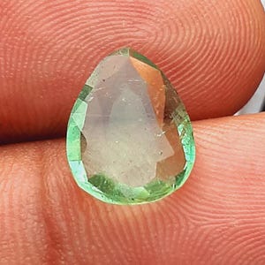 May include: A faceted, pear-shaped gemstone with a light green hue. The gem has multiple facets that catch the light, creating a sparkling effect. The gemstone is held between two fingers, showcasing its size and clarity.