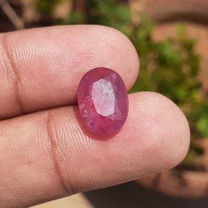 May include: An oval-shaped, faceted gemstone with a deep pinkish-red hue. The stone features a polished surface with multiple geometric facets that reflect light, showcasing a translucent quality and a vibrant, saturated color throughout the gem.