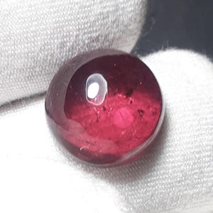 May include: A polished, cabochon-cut gemstone with a deep red hue. The stone has a smooth, rounded shape and a reflective surface, with a bright, circular inclusion near the top. The gem is set against a white background.