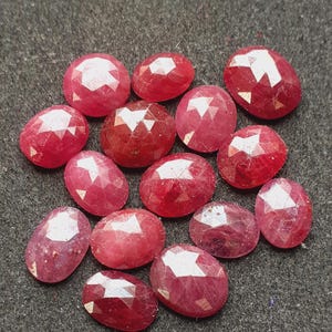 Puede incluir: Un conjunto de gemas de rubí facetadas de forma ovalada. Los rubíes, de color rojo intenso, están dispuestos sobre una superficie gris oscura. Las piedras preciosas tienen una superficie pulida y reflectante.