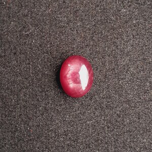 May include: A polished, oval-shaped ruby gemstone. The gem is a deep red color with a reflective surface, set against a dark gray textured background. The stone's smooth, rounded form is highlighted by a bright, white reflection.
