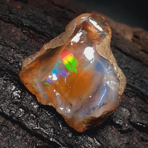 Könnte beinhalten: Ein roher Opal-Edelstein mit einem Farbspiel. Der Stein weist Orange-, Braun- und Weißtöne auf, mit schillernden Blitzen in Grün, Gelb und Blau. Die raue Textur und die natürliche Form des Opals sind sichtbar.