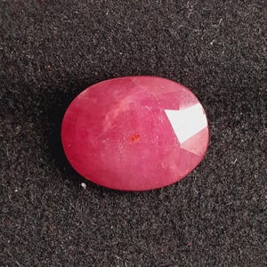 May include: A faceted, oval-shaped ruby gemstone. The gem is a deep, saturated red color with a polished surface. The gemstone is set against a dark, textured background.