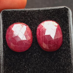 May include: Two oval-shaped, deep red gemstones with multiple facets. The gems are displayed on a black surface. The gems have a brilliant, reflective surface. A finger is partially visible in the upper left corner.