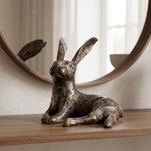May include: A bronze-toned hare figurine with a textured, weathered surface finish. The sculpture depicts a hare in a resting position with its ears pointed upward, showcasing a dark metallic patina with lighter highlights across its form.