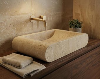 Puede incluir: Un lavabo rectangular de piedra travertino beige con bordes redondeados, que cuenta con un grifo de pared en oro cepillado con agua fluyendo hacia la cubeta. Una toalla de mano beige doblada descansa sobre el mueble de madera oscura.