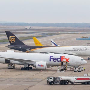 May include: A white FedEx cargo airplane with purple tail branding parked on an airport tarmac next to a brown and white UPS cargo airplane with the text Worldwide Services. A yellow DHL airplane and a white fuel tanker truck are also visible.