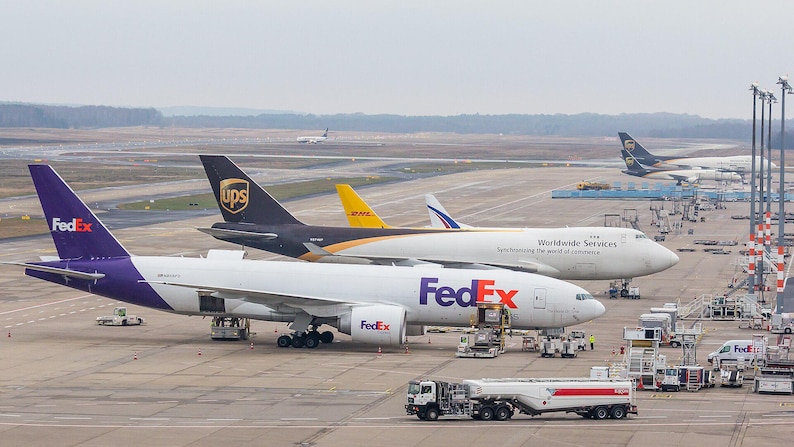 May include: A white FedEx cargo airplane with purple tail branding parked on an airport tarmac next to a brown and white UPS cargo airplane with the text Worldwide Services. A yellow DHL airplane and a white fuel tanker truck are also visible.