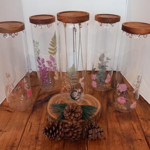 Könnte beinhalten: Fünf transparente Glas-Ausstellungsgläser mit Holzdeckeln, jedes mit zarten floralen und botanischen Designs verziert. Die Gläser sind auf einer Holzoberfläche angeordnet, mit einer dekorativen Holzscheibe und Tannenzapfen im Vordergrund.