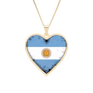 Può includere: Una collana con pendente a forma di cuore color oro. Il pendente presenta la bandiera argentina, con sfondo azzurro, una striscia bianca e un emblema del sole. Anche la catena della collana è color oro.