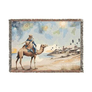 Könnte beinhalten: Gewebte Wurfadecke mit einer Aquarellillustration einer Person, die auf einem Kamel auf ein Dorf am Strand zureitet. Die Szene spielt unter einem Sternenhimmel mit hellem Licht. Die Decke hat einen Fransenrand.