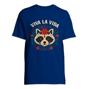 May include: A royal blue t-shirt featuring a raccoon design with a floral wreath and the text "VIVA LA VIDA" in a decorative font. The raccoon has a sugar skull-inspired design with red flowers and accents. The shirt is a short-sleeved crew neck.