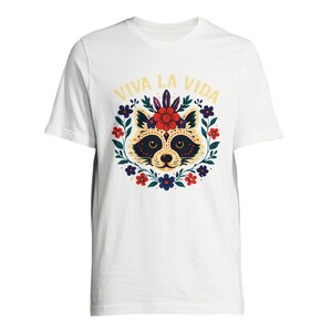 May include: White t-shirt featuring a raccoon face design with a floral wreath and the text "VIVA LA VIDA" in yellow. The raccoon has a decorative face with a flower crown. The design is centered on the shirt.
