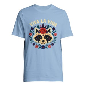 May include: Light blue t-shirt featuring a raccoon face design surrounded by a floral wreath. The raccoon has a black mask and is decorated with flowers. The text "VIVA LA VIDA" is arched above the raccoon.