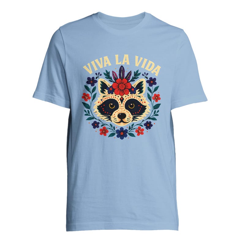 May include: Light blue t-shirt featuring a raccoon face design surrounded by a floral wreath. The raccoon has a black mask and is decorated with flowers. The text "VIVA LA VIDA" is arched above the raccoon.