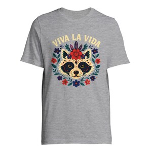 May include: A gray t-shirt featuring a raccoon design with floral accents and the text "VIVA LA VIDA" in yellow. The raccoon has a decorative face with a floral crown, surrounded by red, blue, and purple flowers.