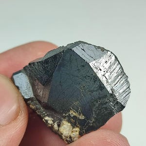 Könnte beinhalten: Dunkles, facettiertes Mineralexemplar mit metallischem Glanz. Der Kristall ist überwiegend schwarz mit reflektierenden silbergrauen Oberflächen. Die Basis des Minerals hat eine raue, beige und braune Textur. In einer Hand gehalten.