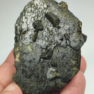 Op de afbeelding: Een donkergroen mineraal specimen met een ruw, getextureerd oppervlak. Verschillende kleine, donkere, langwerpige kristallen zijn ingebed in de matrix. Het mineraal heeft op sommige plaatsen een metallic glans en het algemene uiterlijk is ruig en natuurlijk.