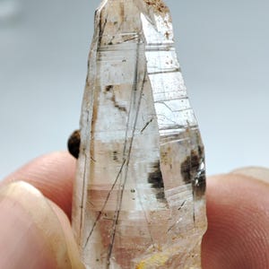 May include: A clear quartz crystal with dark inclusions and a pointed tip. The crystal is held in a hand, showcasing its natural, raw form. The quartz has a translucent quality, with visible internal structures and a slightly rough texture.
