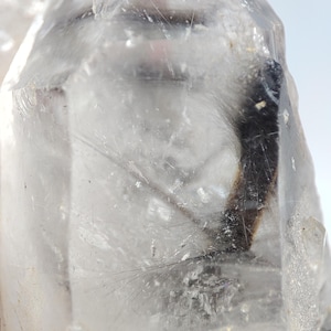 May include: Close-up of a clear quartz crystal with internal inclusions. The crystal is transparent with visible lines and a dark, elongated inclusion running through the center. The overall shape is a natural, pointed form.