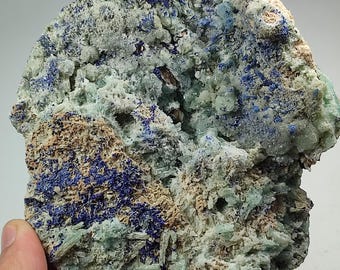May include: A rough-hewn mineral specimen featuring a mix of colors. The rock has a light green base with patches of deep blue and areas of tan and brown. The texture is rough and crystalline, with visible formations.