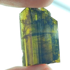 May include: A translucent gemstone, held between fingers, displaying a blend of yellow, green, and blue colours. The crystal has a rectangular shape with a faceted top. The natural, raw form of the gemstone is visible.