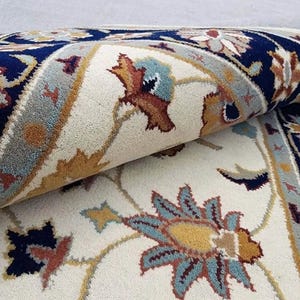 May include: Rolled-up area rug with a cream-colored background and a detailed floral pattern. The design incorporates navy blue, light blue, gold, and rust-colored accents. The rug's border features a repeating geometric pattern.